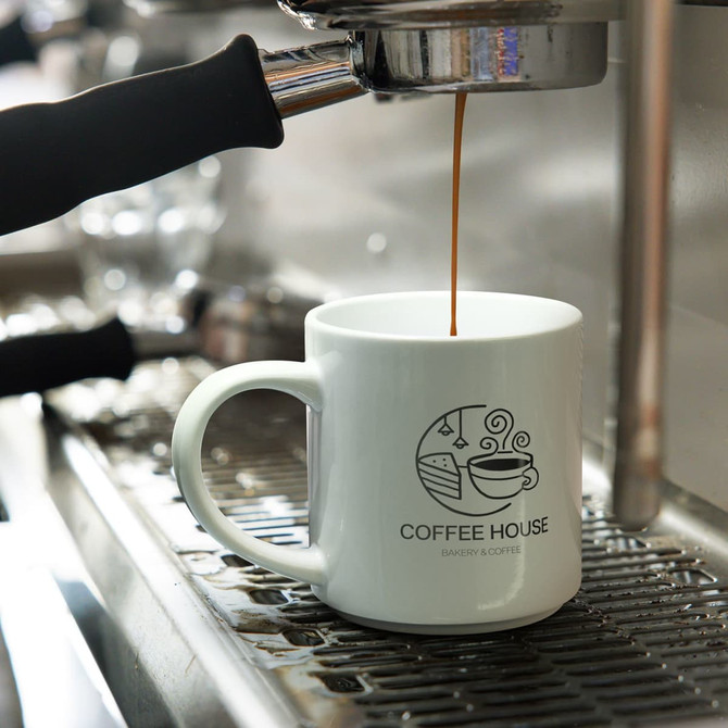 A stackable white coffee mug with a logo, placed under an espresso machine as coffee is being poured.