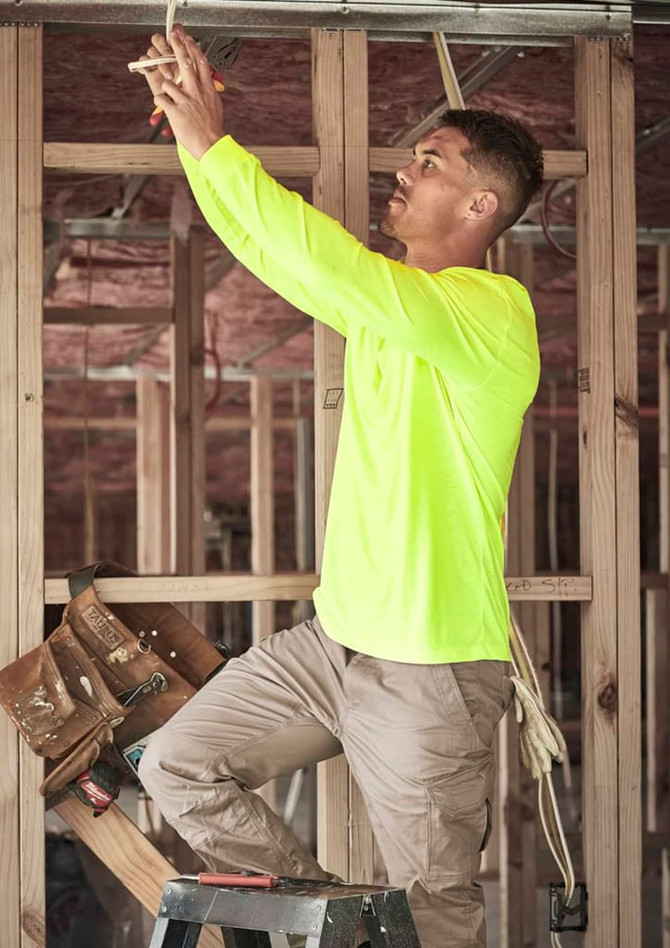 A man in a high visibility long sleeve yellow tee works on a ladder in a construction setting. The shirt has a logo.