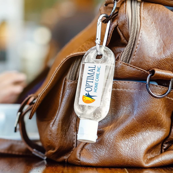 A clear 30ml hand sanitiser with a white cap, attached to a brown bag via a carabiner. Features a logo.