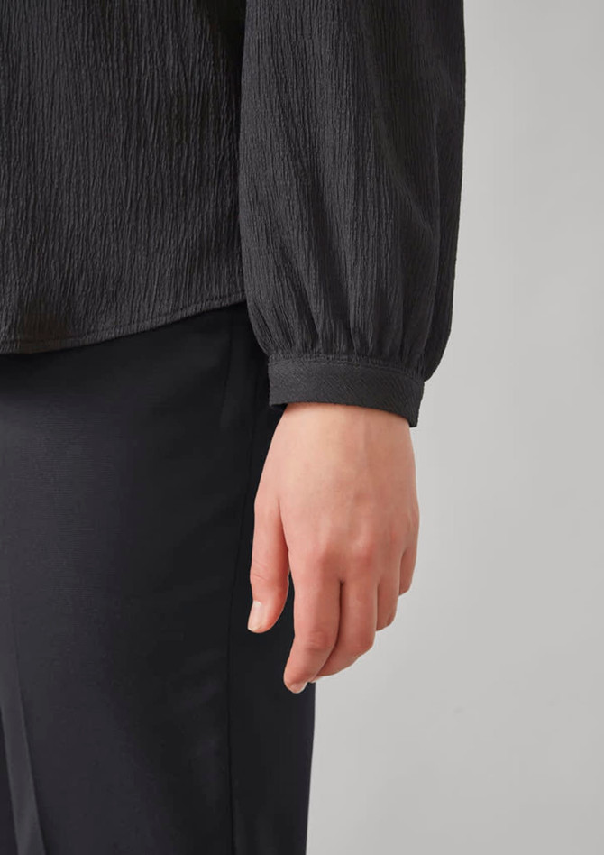 A black long sleeve blouse featuring a ruched texture and gathered cuffs, paired with black pants.
