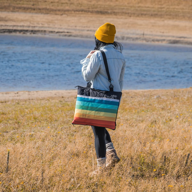 A person wearing a yellow beanie carries a multi-coloured striped picnic blanket against a natural landscape.