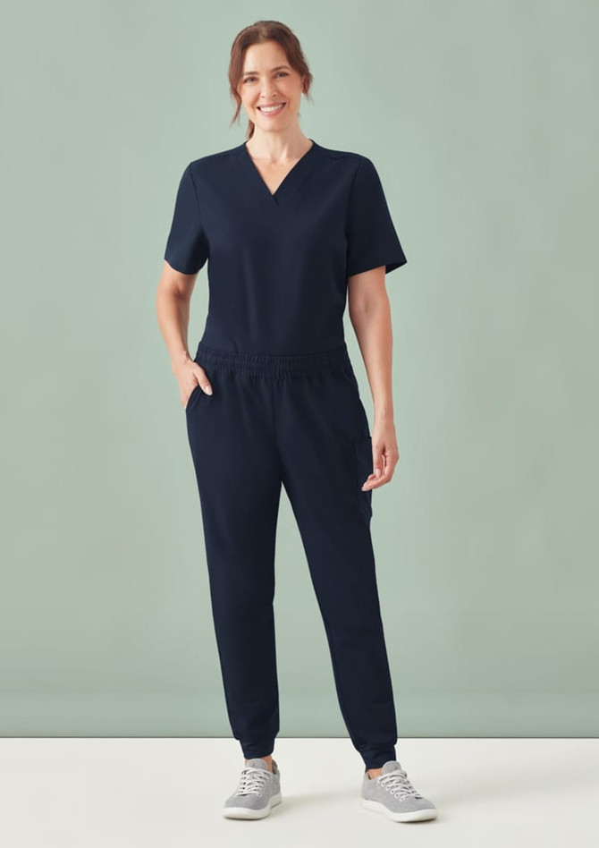 A navy women's scrub top with a V-neck and short sleeves, paired with matching pants, modelled by a smiling woman.