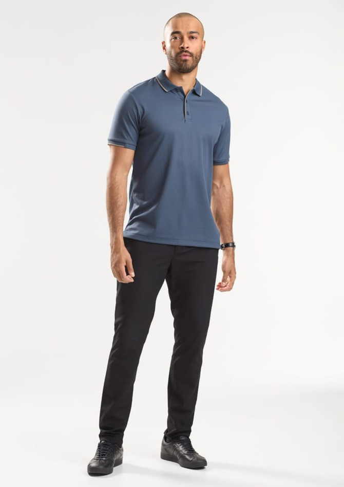 A man wearing a blue short sleeve polo shirt and black trousers, standing against a light background.
