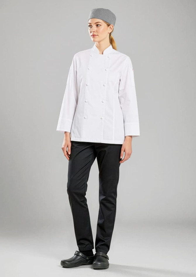 A woman in a white long sleeve chef jacket, black pants, and a grey chef's hat, standing against a grey background.