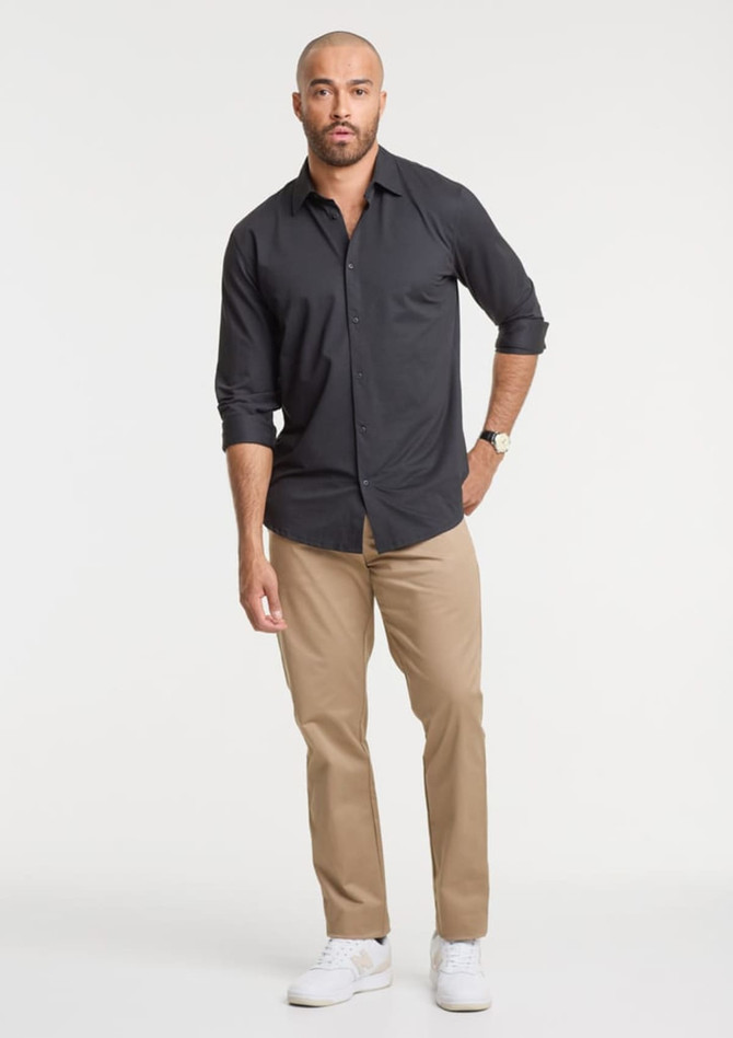 A man wearing a black long sleeve shirt and beige trousers, standing with hands at his sides and wearing white shoes.