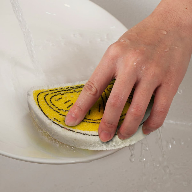 A yellow and white expandable sponge is being held under running water, with droplets visible on its surface.