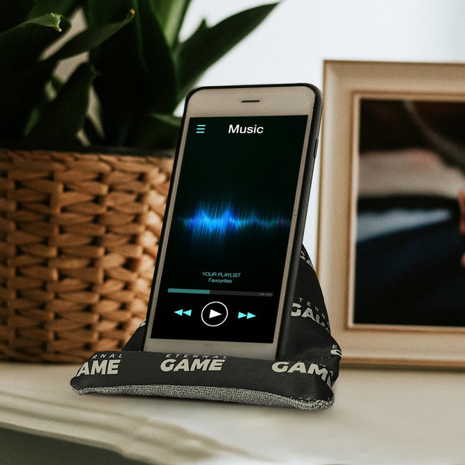 A black bean bag phone holder supports a smartphone displaying a music app, set against a decorative background.