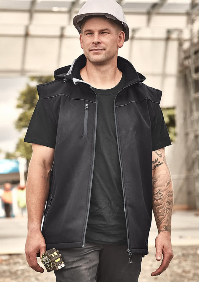 A unisex black softshell jacket features a hood, zippered pockets, and a branding logo, worn by a man at a construction site.