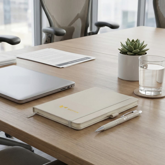 A tan notebook and a cream pen on a wooden table, accompanied by a laptop, a glass of water, and a small plant.