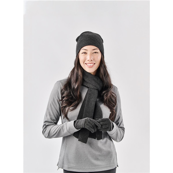 A woman wearing a black knit beanie, long black scarf, and grey long-sleeve top, smiling against a plain background.