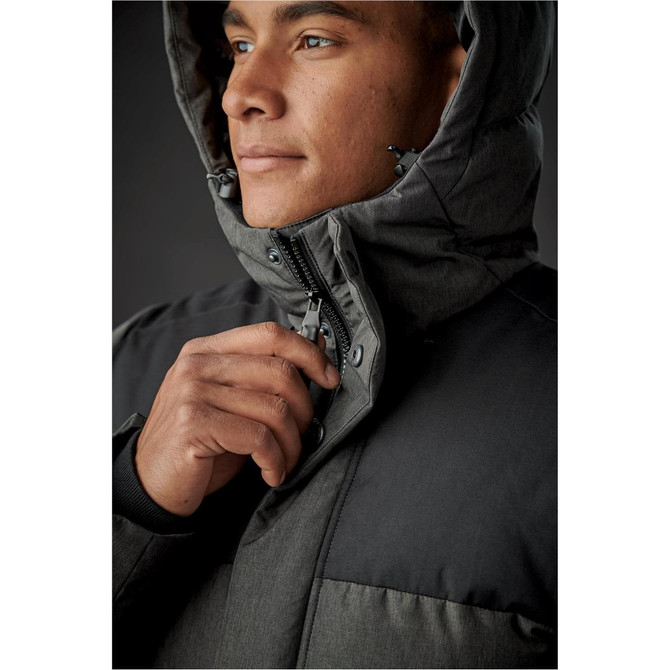 A close-up of a man in a grey and black parka jacket, partially zipped, with the hood up. The jacket features a logo.