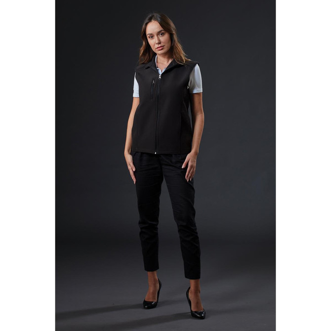 A black softshell vest is worn by a woman styled with sleek trousers and heels against a dark background. It features a logo.