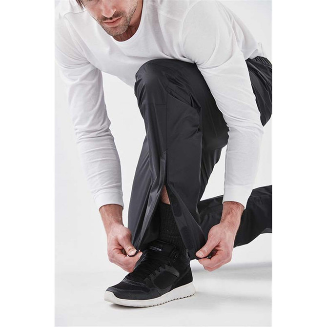Men's Olympia Rain Pant in black, featuring a zip detail at the ankle. Worn by a model kneeling and adjusting the pant.