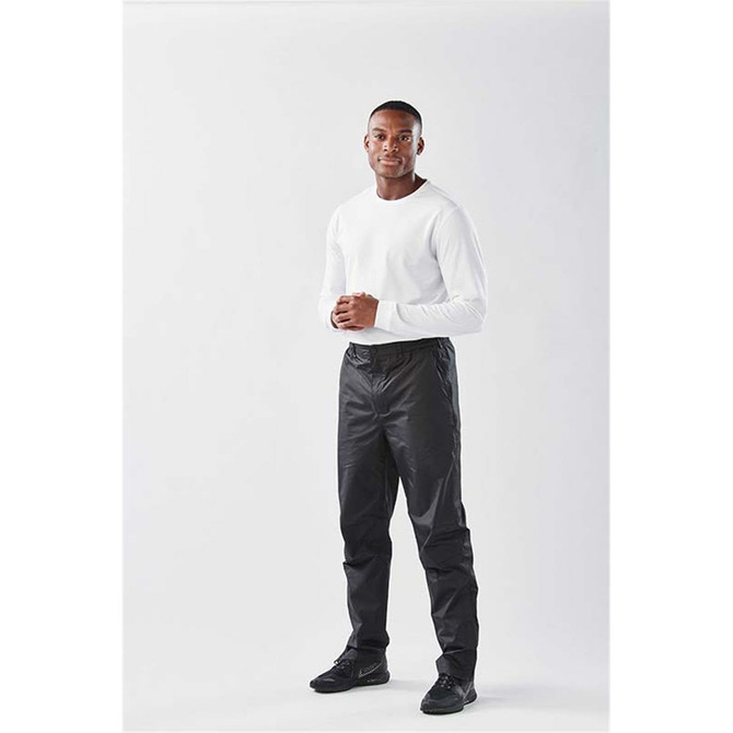 A man stands wearing black rain pants and a white long-sleeve shirt. The pants have a logo on them.