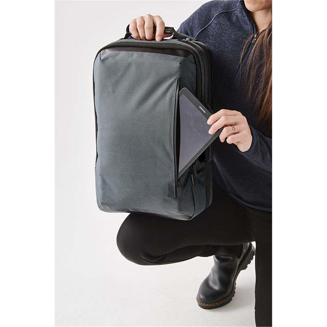 A person holds a grey backpack while placing a tablet into a front pocket. The backpack has a logo.
