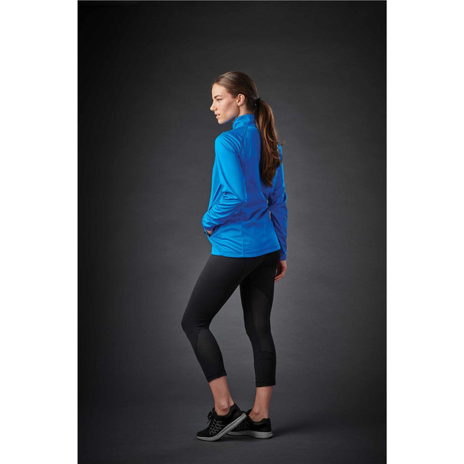 A woman stands sideways wearing a blue lightweight softshell jacket and black leggings, set against a dark background.