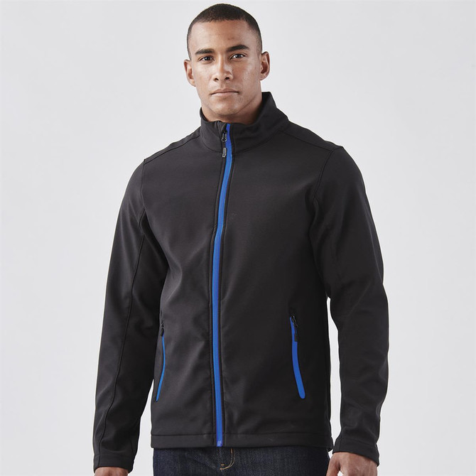 A man wearing a black softshell jacket with blue accents and zippered pockets, featuring a logo.