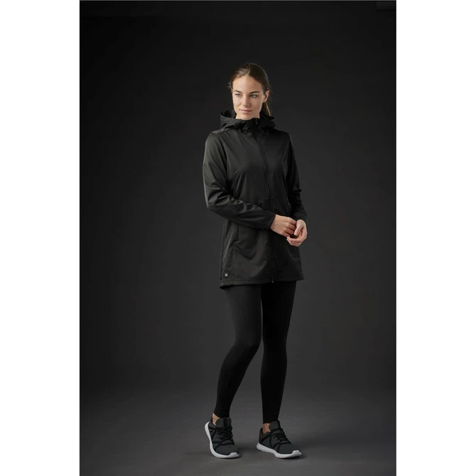 A woman in a black softshell jacket stands against a dark background, paired with black leggings and shoes.