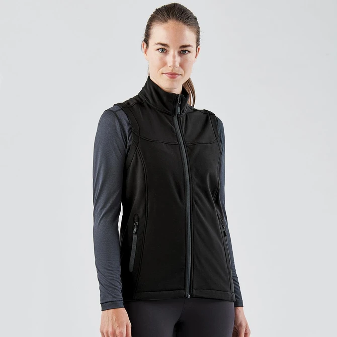 A black softshell vest with a zip front and side pockets, worn over a long-sleeve top. The model has a neutral expression.