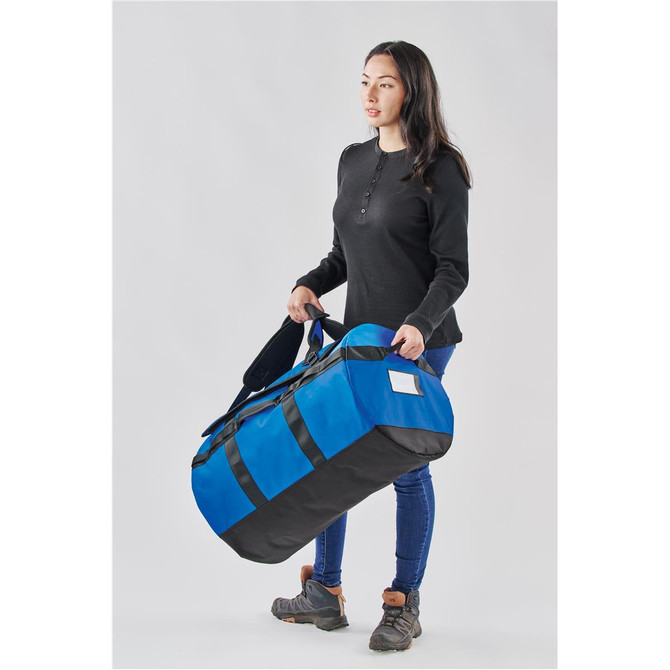 A woman holds a large blue and black duffle bag with straps. The bag features a logo.