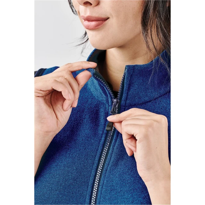 A close-up of a woman in a blue vest, holding the zipper with both hands. The vest has a branding logo.