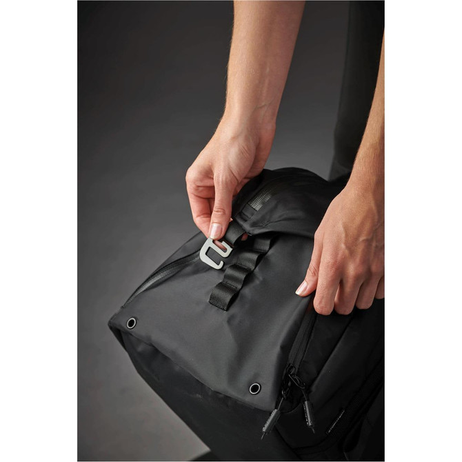 A black duffle bag being held, with a hand fastening a strap on the top. The bag features a logo.