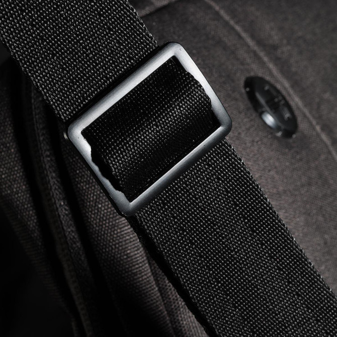 A close-up of a black strap buckle on a bag, featuring a textured fabric and a circular logo.