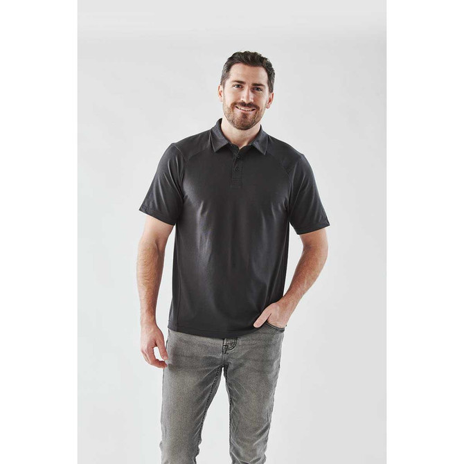 A man wearing a black short sleeve polo shirt, paired with grey jeans, standing against a plain background.