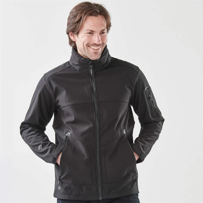 A dark black softshell jacket with a full zip, featuring side pockets and a collar, worn by a smiling man.