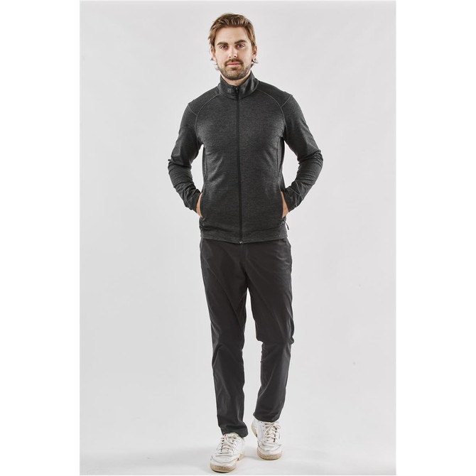A man wearing a black performance jacket with a zip front and side pockets, paired with dark trousers and sneakers.