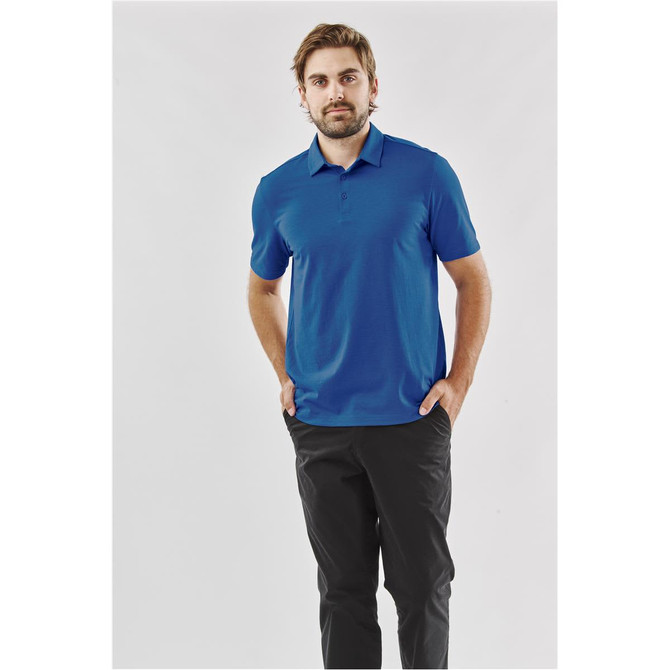 A man wearing a blue polo shirt stands against a plain background, paired with black trousers. The shirt has a logo.