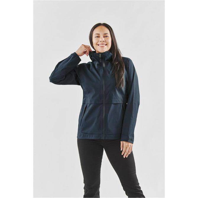 A woman wearing a navy blue technical shell jacket with a zip-up front and high collar, featuring a logo.