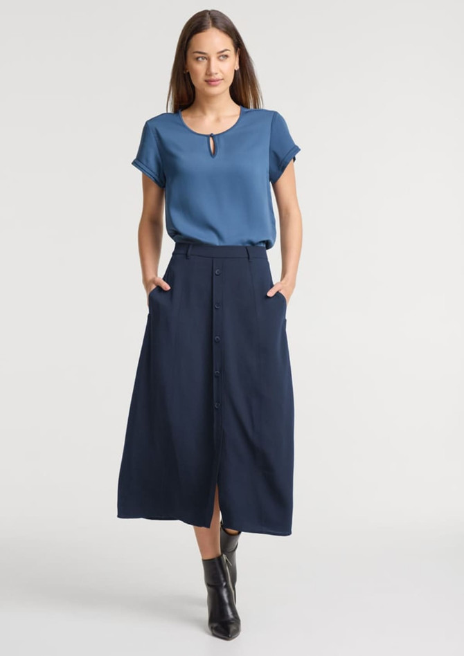 A woman wears a light blue top and a long navy skirt with buttons and pockets. The outfit is stylish and modern.