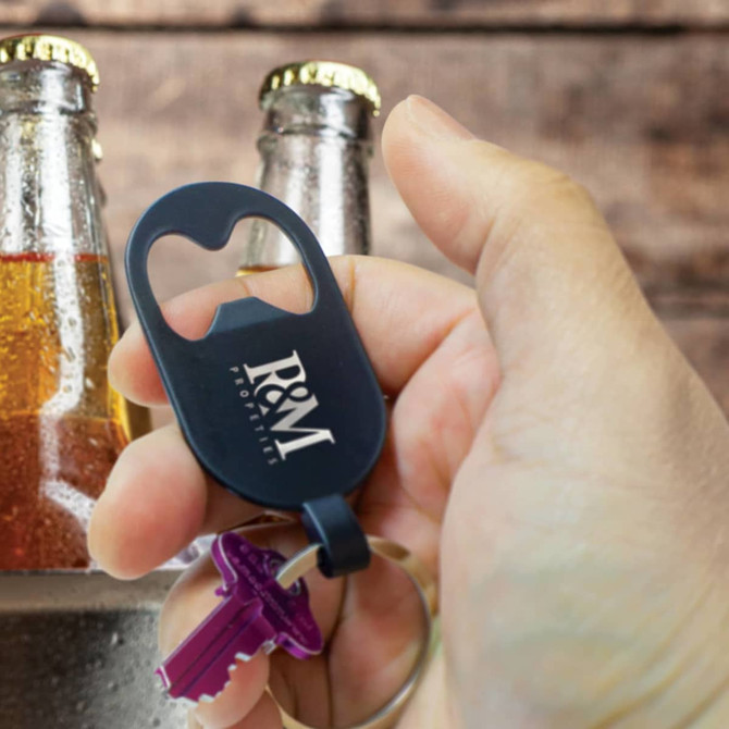 A drink bottle opener key ring in black, held in a hand, with glass bottles in the background. It features a logo.