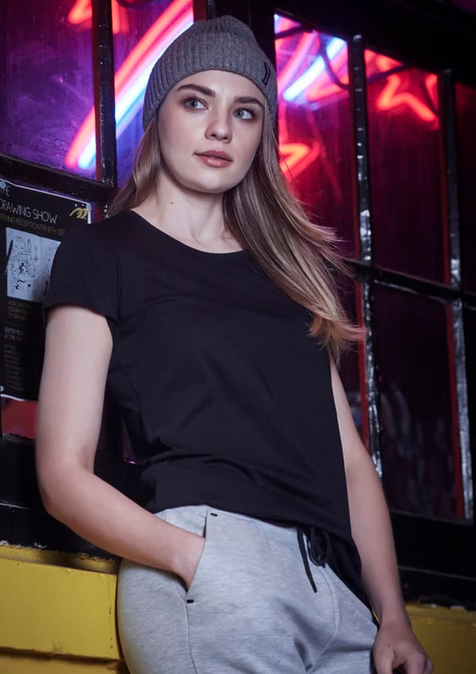 A woman in a black t-shirt and grey pants leans against a window, wearing a grey beanie. The background features neon lights.