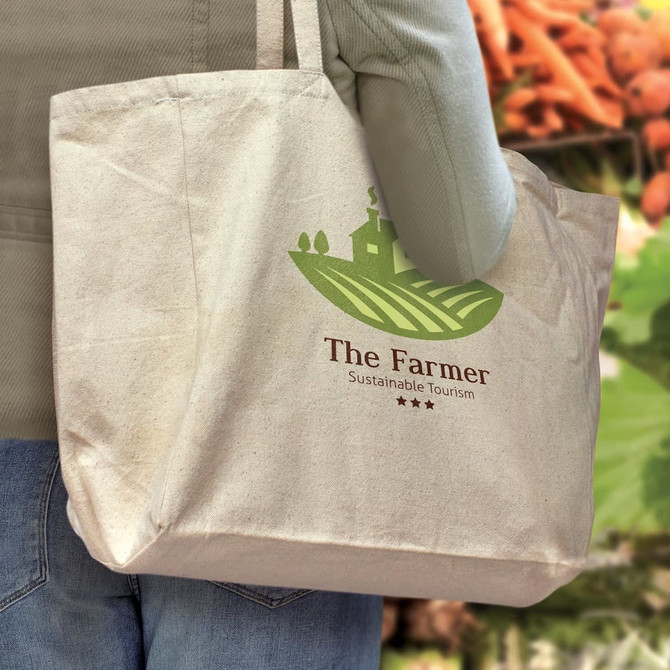 A natural cotton tote bag with a logo, held by a person against a blurred background of vegetables.