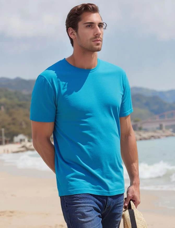 A blue t-shirt worn by a man standing on a beach, with mountains in the background and a straw hat in his hands.