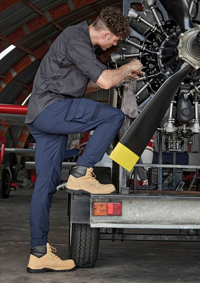 A man wearing navy cuffed work pants and tan boots is performing maintenance on an aircraft engine.
