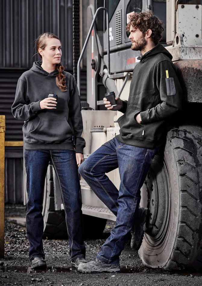A man and woman wear black hoodies and blue denim work jeans, standing beside a large vehicle in an industrial setting.