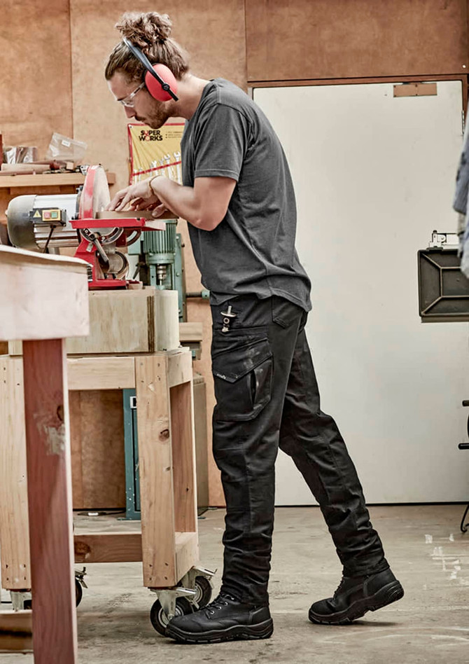 Men's rugged cooling stretch pants in black, featuring cargo pockets, are worn by a person working in a workshop.