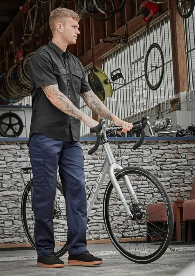 A man in a black shirt stands with a bike, wearing navy midweight drill cargo pants in a workshop setting.