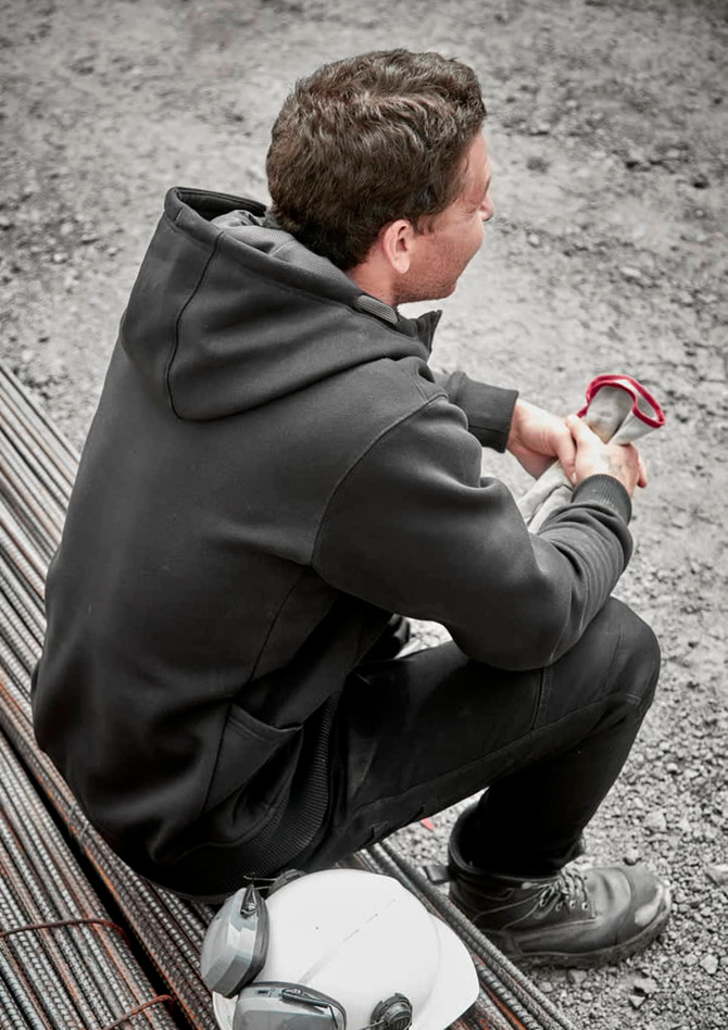A unisex zip front hoodie in black, featuring multiple pockets, worn by a seated person on a construction site.