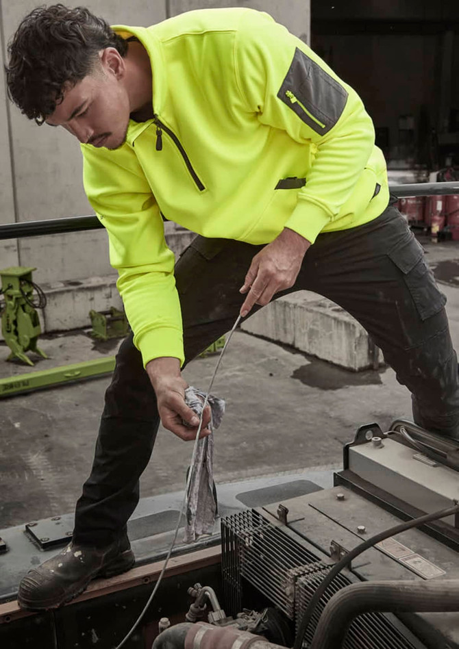 A man wearing a bright yellow water-resistant 1/4 zip pullover is working on machinery. The pullover has a logo.