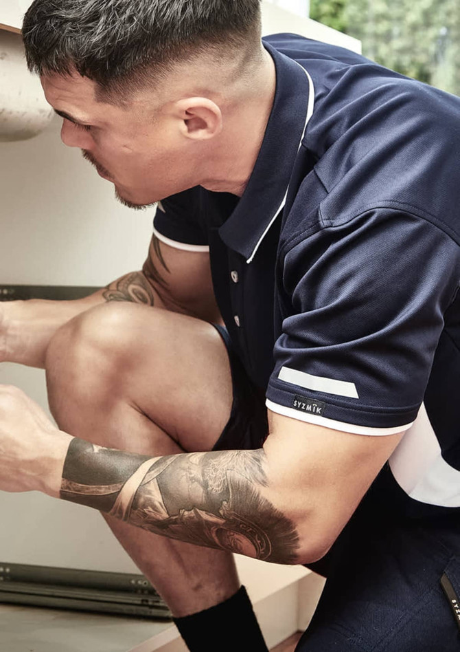 A man wears a navy and white short sleeve polo shirt while crouching, showcasing his tattooed arm and a logo on the sleeve.