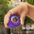 A hand holds a purple wooden yoyo featuring a logo. The background shows a blurred outdoor scene.