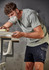 A man in a light green T-shirt and dark shorts, wearing a face mask, works at a table. The shirt has a logo.