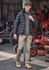A black hooded puffer jacket is worn over a grey shirt, paired with beige pants and tan boots in an outdoor setting.