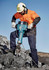 A man in safety gear operates a jackhammer on a rocky surface, wearing an orange shirt and navy blue cargo pants.