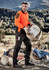 A man in an orange shirt and black tough work pants carries a bucket at a construction site, with mountains in the background.