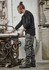 A man wearing black work pants stands by a workshop table, handling tools in a well-equipped space.